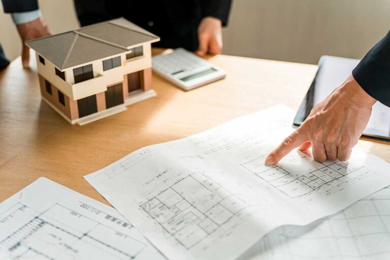 Home plans, a site plan, and a hard hat arranged on a construction desk beside a partially built house.
